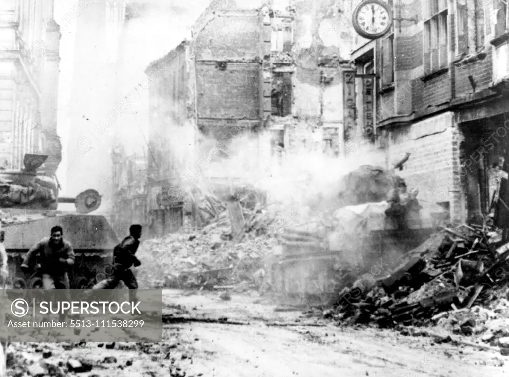 Dramatic Picture of the Battle For Cologne: During fierce fighting in the shadow of the Cologne Cathedral, a U.S. tankman, one leg blown off, lies on top of his was machine after he was blown from the turret by a German shell. While one First U.S. Army soldier (right) rushes to aid the wounded soldier, another runs for stretcher bearers. The wounded solider was the only members of the tank crew to survive the shell hit. The rubble-strewn street is the Komoodientrasse, running from Cathedral Squa