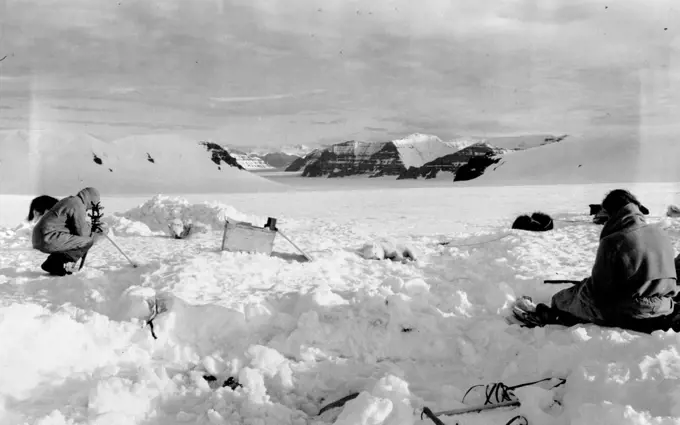 British Arctic Expedition: View of new mountains. Godfrey on left taking a theodolite observation with Lindsay "booking" for him. Hollow in foreground where tent had been pitched. The flat basalt formation of the mountains. November 26, 1934. (Photo by Martin Lindsay).