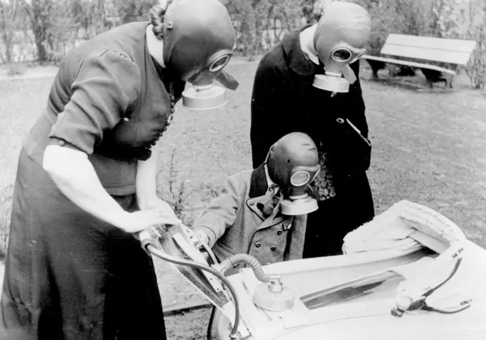 German Families Are All "Gas Proof" Now! -- A baby in his gas proof cradle after he had been placed in his pram, with his mother, operation the bellows air pump placed on the pram handles, little brother and aunt, all wearing gas masks, looking on. As in France and Great Britain, gas masks have now been issued to all German Families. The designs of masks are of difference types, Suitable for adults, children, and babies, the last-named being placed in a kind of gas proof cradle, operated by a b