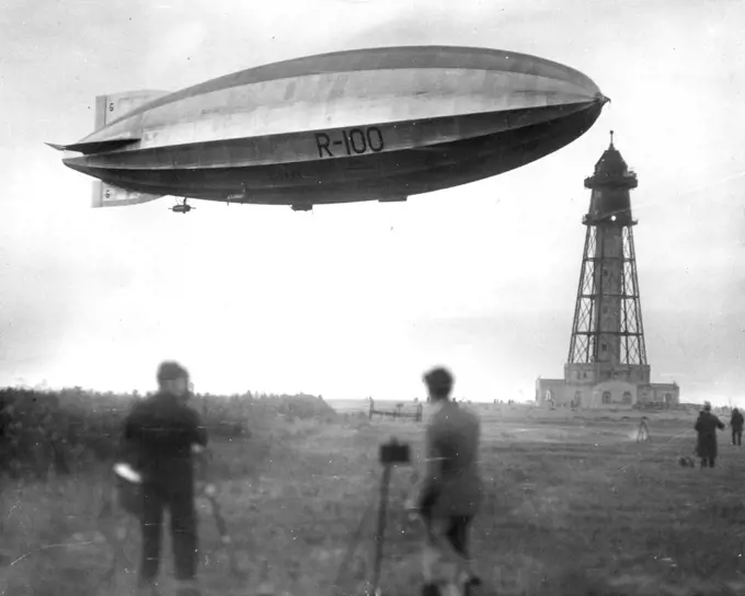 A Giant Visitor -- From Out Of The Clouds: Greeted by the wild cheers of enthusiastic spectators and the whir of airplane propellers, the stately R-100, Newest dirigible king of the Atlantic, Gracefully points its nose toward the new $1,000,000 Mooring-Mast especially constructed in her new world. This view, taken from the ground, shows the giant visitor just before actual contact with the mast had been established by means of the land and aerial cables which had been hocked together. January 8,