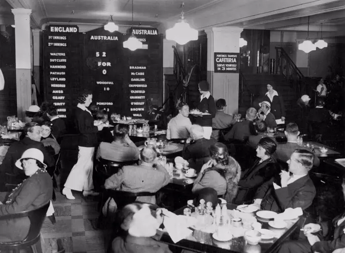 Lunch Scores -- Diners watching the scoreboard in operation today. Note waitresses in white trousers. A Cricket scoreboard has been created in the restaurant of Messrs. Pontings' Stores, so that the diners in London may watch the progress of the test match, run by run. Watch run is recorded immediately it is scored. June 8, 1934. (Photo by London News Agency Photos Ltd.).