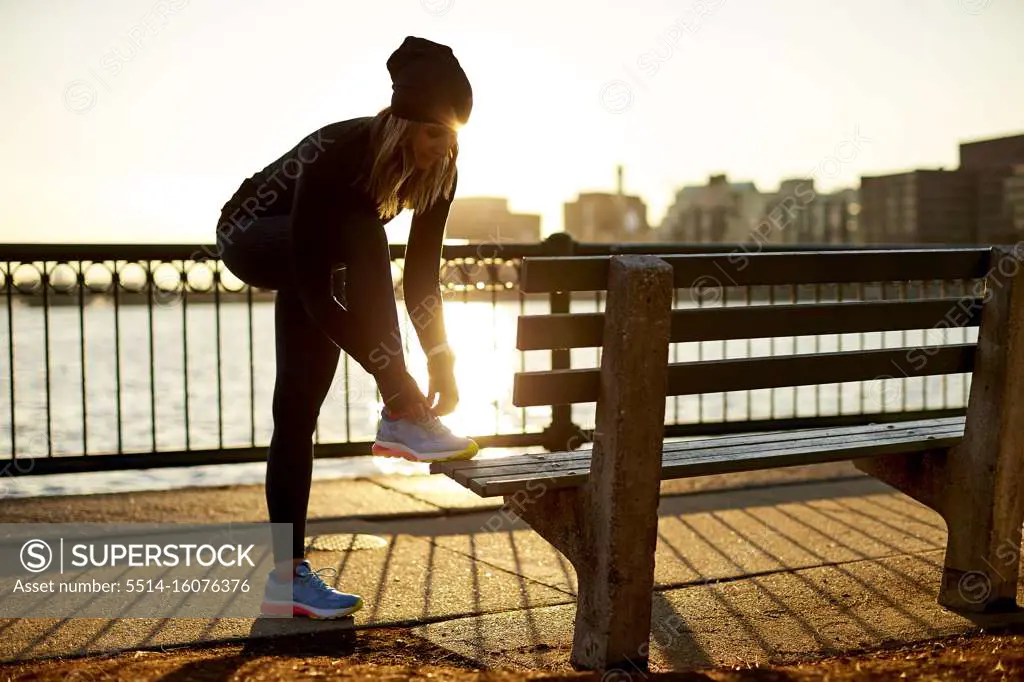 A backlit athletic woman ties her sneaker on a park bench.