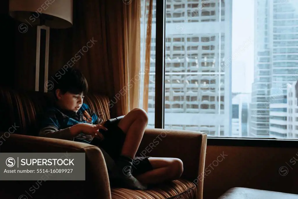 Boy using tablet in chair beside window with tall buildings outside.