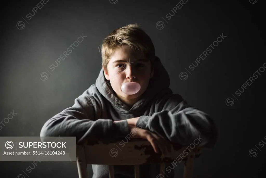 Teen boy in hoodie sitting on a chair in dark room blowing a bubble.