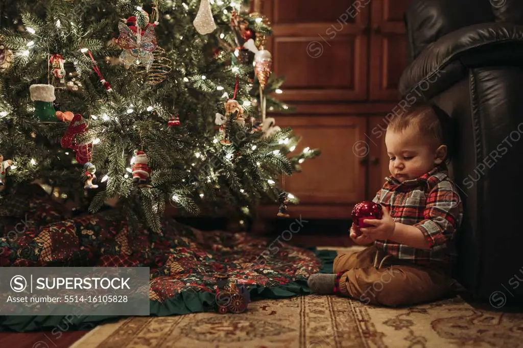 Toddler boy sitting under Christmas tree holding ornament