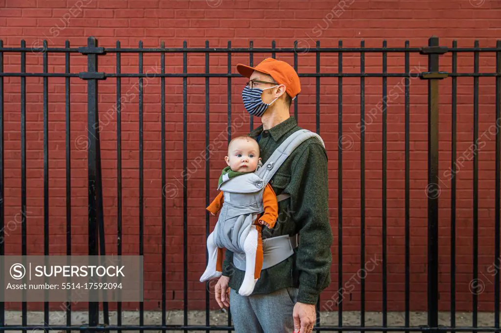 Father wearing face mask carrying cute daughter in baby carrier while standing by fence during coronavirus crisis