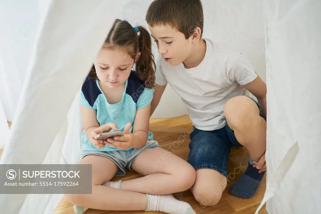 Boy and girl looking at a smartphone inside a white teepee tent inside their house. Technology concept