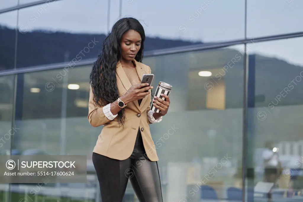Portrait of black woman wearing a brown suit talking on the phone and holding a container with coffee in the street