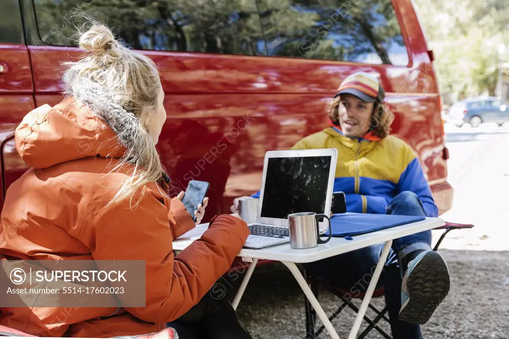 Young couple working outside their red campervan with a forest