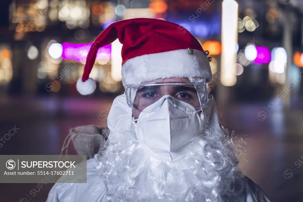 Portrait of a man, with the hat and beard of Santa Claus, but with a face mask, glasses and coverall, to protect himself from Covid 19. In the background the empty city.