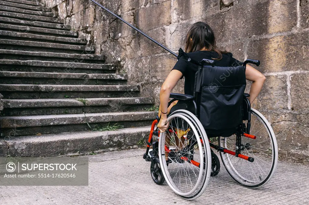 Rear view of a young woman in a wheelchair looking at some stairs. Con