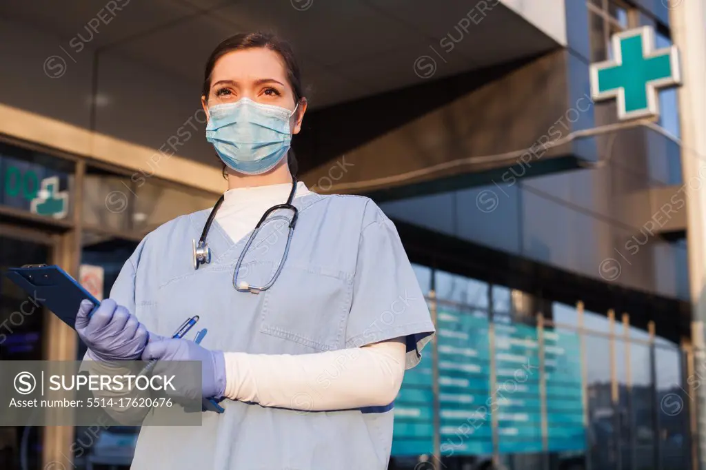 Young female doctor standing in front of healthcare facility, wearing protective face mask and PPE equipment, holding medical patient clipboard, COVID-19 pandemic crisis