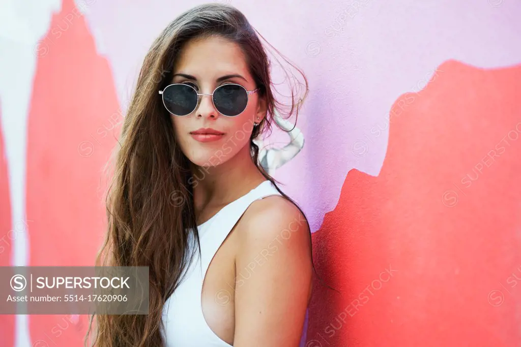 Young woman with long hair wearing stylish sunglasses and standing near red building wall in city