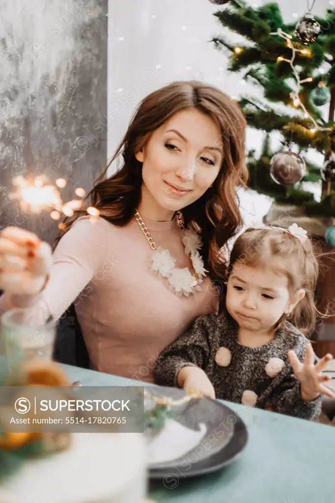 Young family with a daughter in festive outfits at a served table with candles, garlands, sparklers and a cake near the Christmas tree on New Years Eve