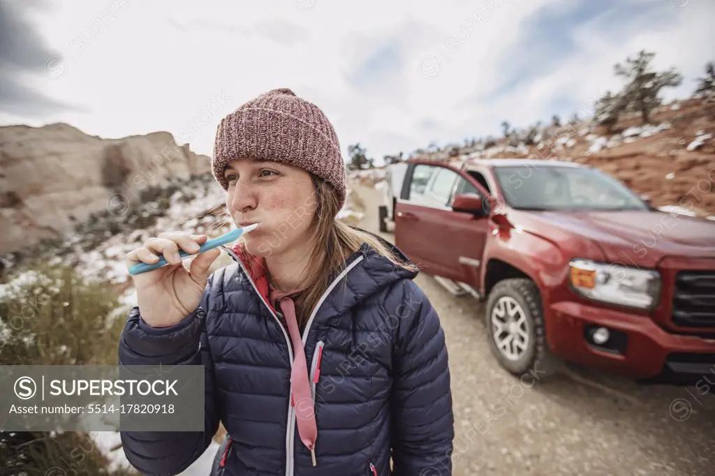 Woman in jacket and hat brushes teeth while car camping in Utah desert