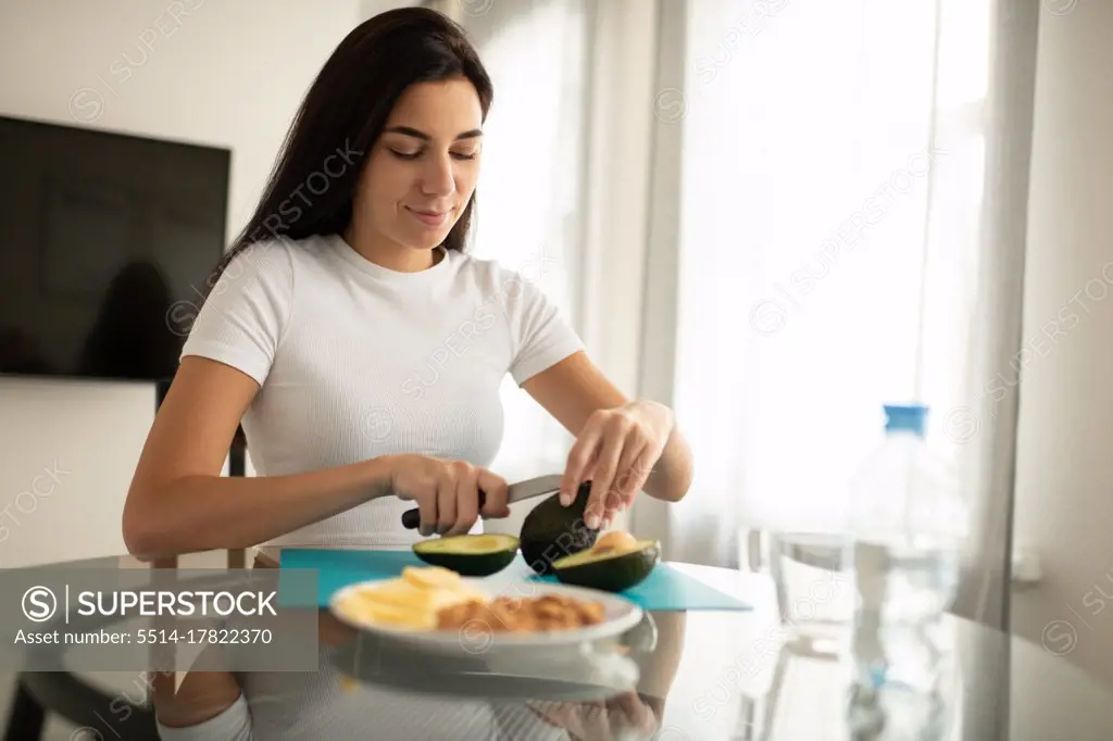 Content female preparing snack of avocado sitting in kitchen