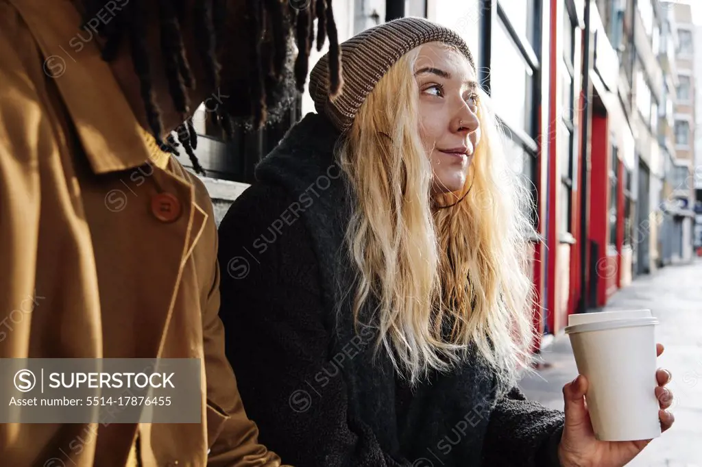 blonde young female sitting outside while drinking coffee with her friend. multicultural friendship