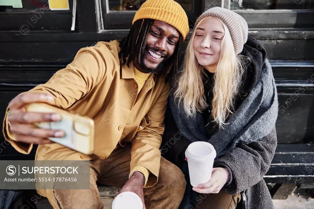 cool biracial friends taking a selfie while sitting outside holding their cup of coffee