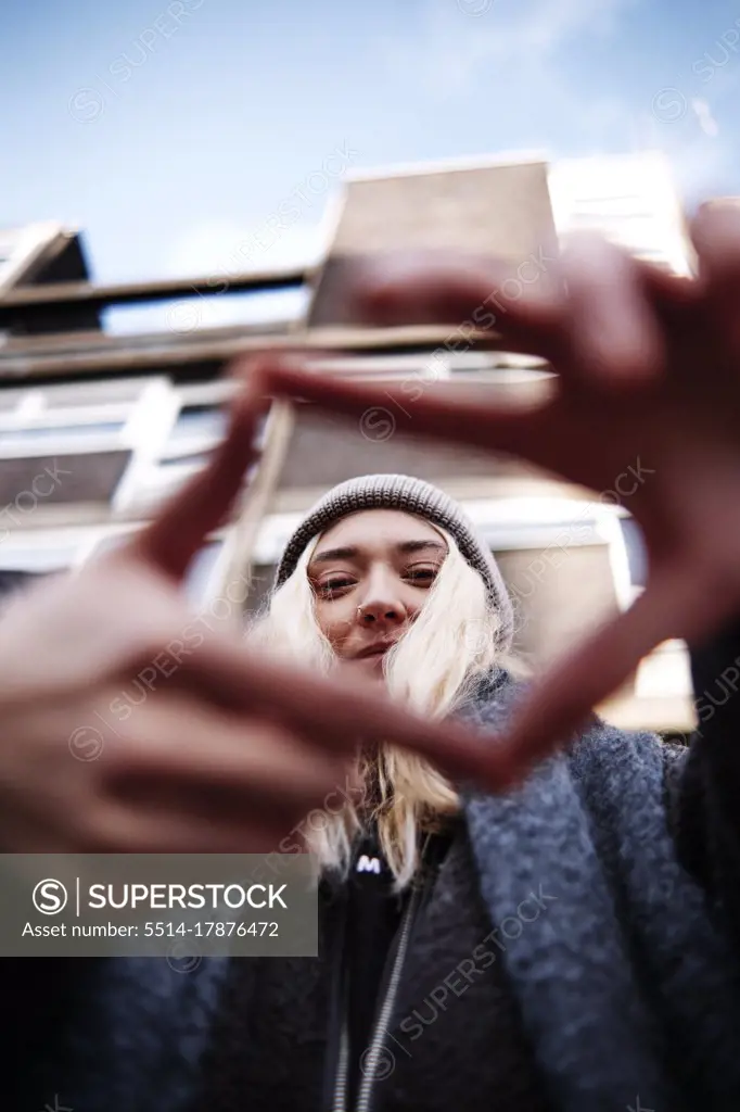 vertical shot of young female looking at camera, smiling through finger frame in front of a building. low angle shot.