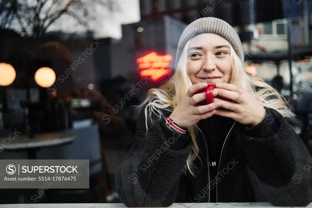 young female drinking coffee, looking through a window with reflection of the city