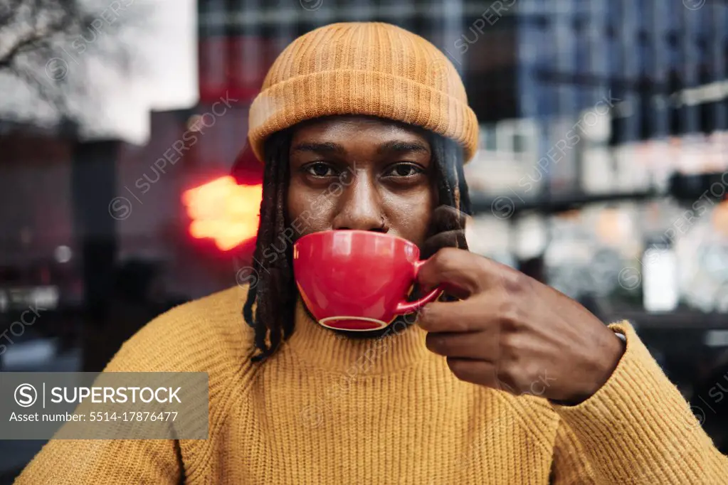 young african guy staring at camera as he drinks a cup of coffee at a cafe, reflection of the city on window