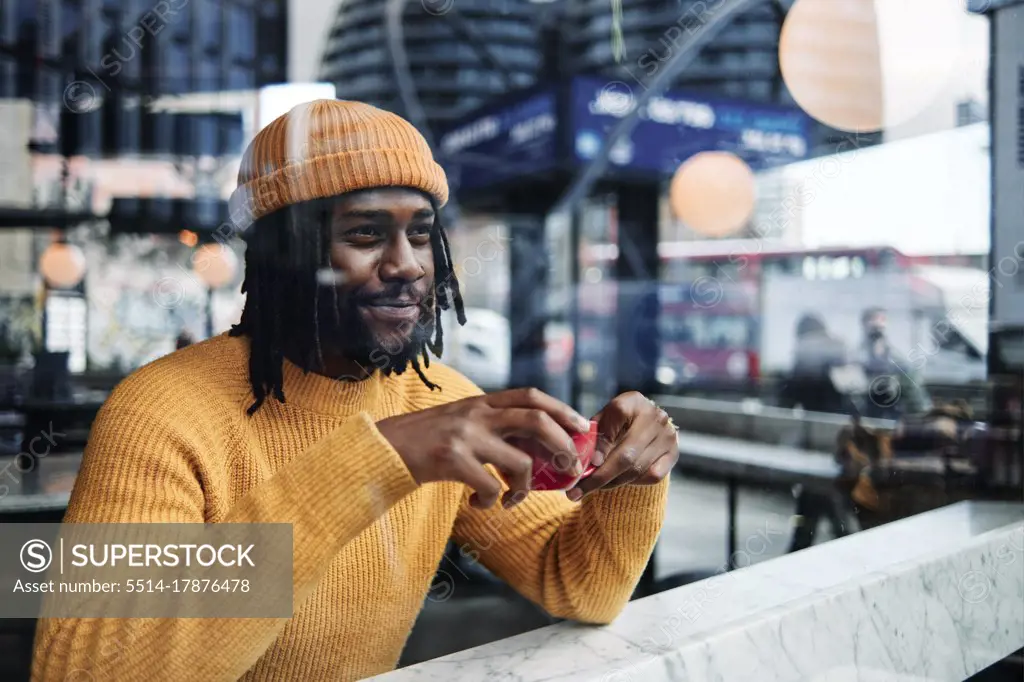 young african guy looking through a window as he drinks a cup of coffee at a cafe, reflection of the city on the glass