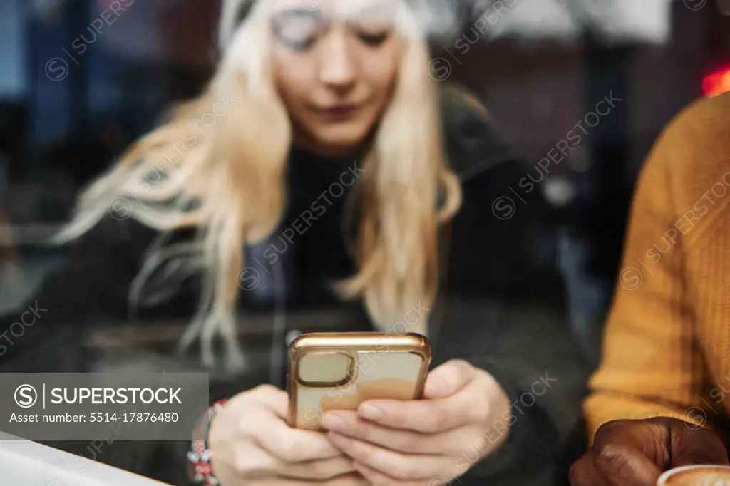 young girl checking her phone, sitting at the window of a cafe. reflection on the glass. focus on the phone