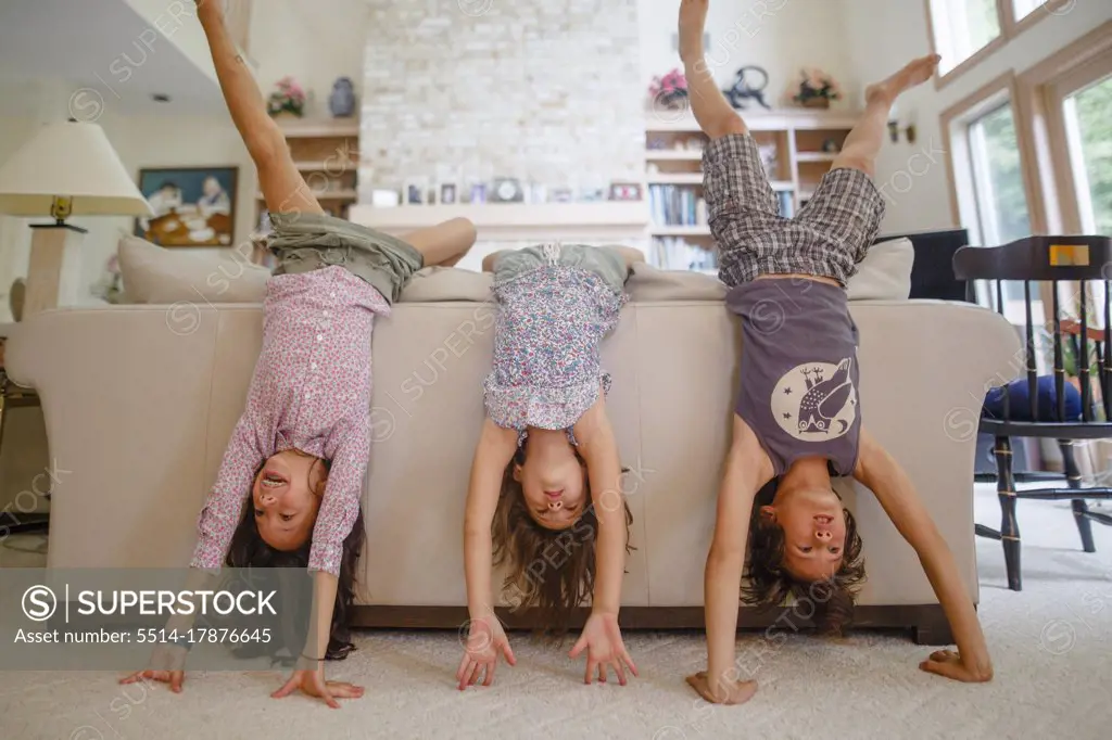 Three children lean against couch in living room doing handstands