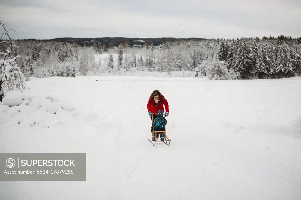 Mother pushing child on sleigh in snow in woods on cold winter day