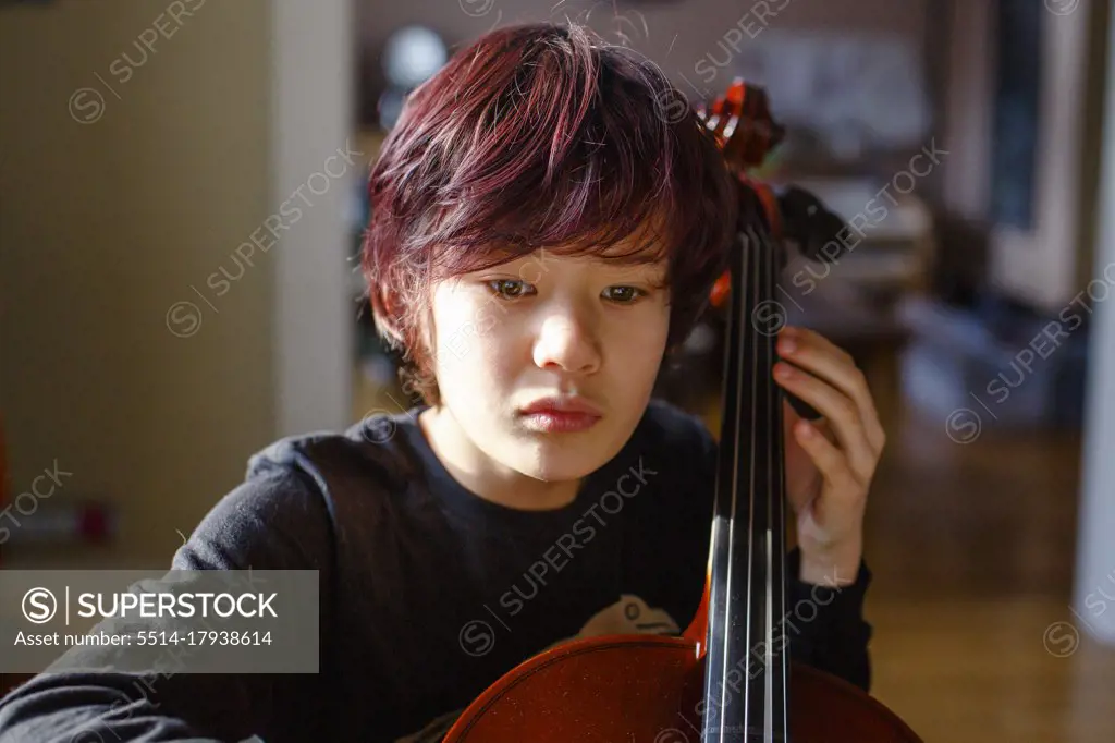 A boy with intense focus and dyed red hair plays cello in window light