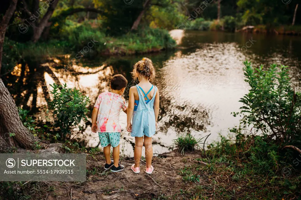 Two young kids standing at edge of lake pointing at fish in the water