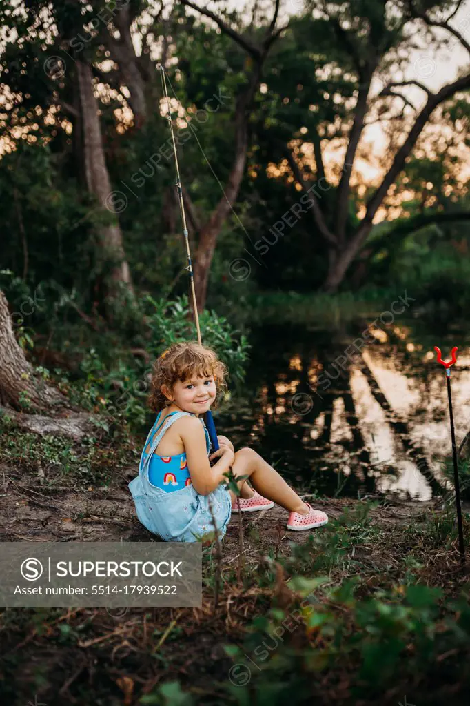 Young girl sitting at creek bank holding fishing pole and smiling