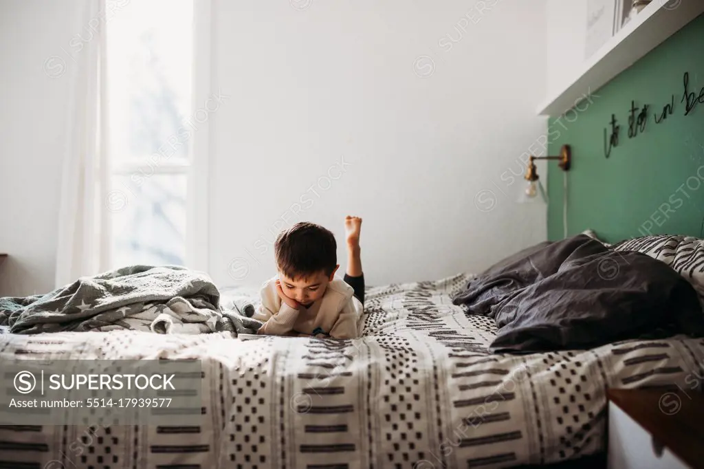Young boy laying on bed watching on tablet in modern bedroom