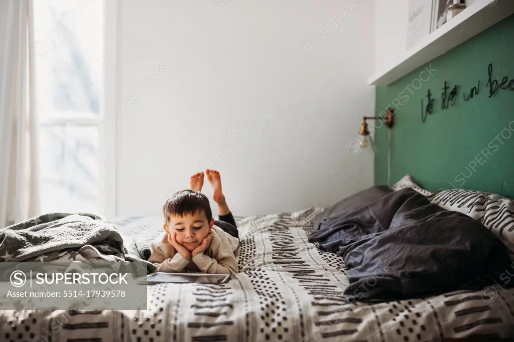 Happy young boy watching videos on tablet while laying on large bed