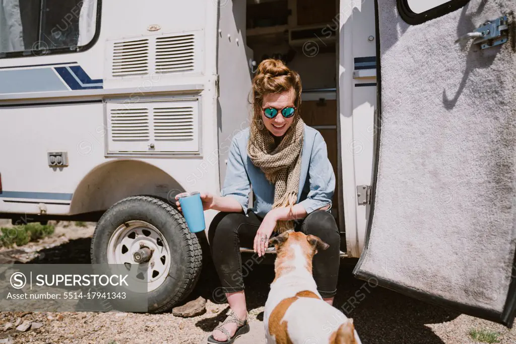 A stylish young woman and her dog sit outside their trailer in the sun