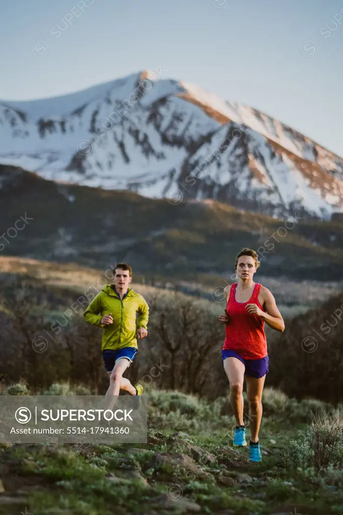 A woman and man trail run at sunset with snowy peak in the distance