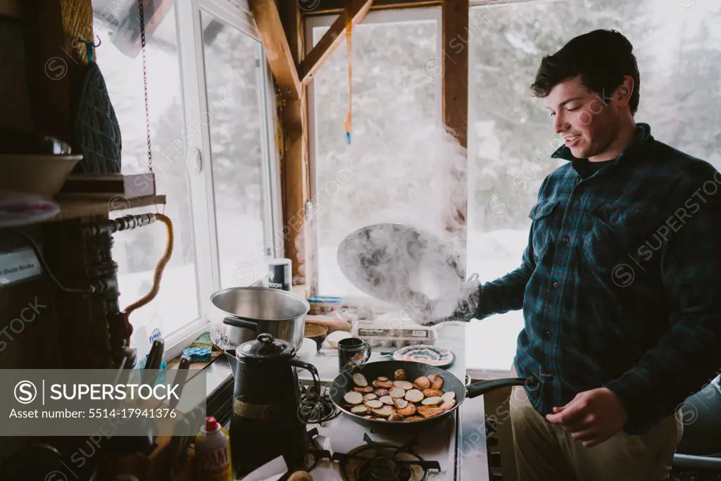 A young man cooks steaming breakfast in a cabin during winter