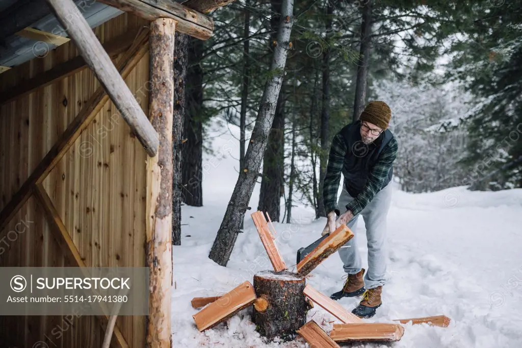 A man wearing flannel shirt chops firewood outside in the snow