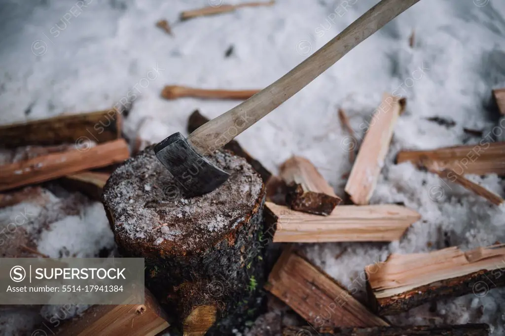 A wood handled axe sticks into a log with firewood and snow around it