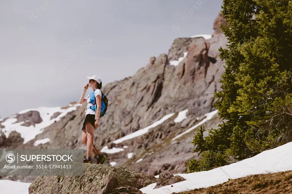 Woman trail runner sips water in front of pine trees snow and rocks