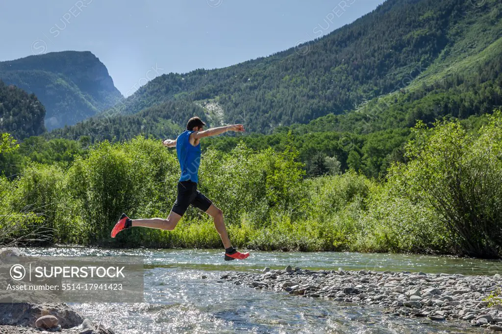 Male trail runner leaps over rocky river on sunny day in the mountains