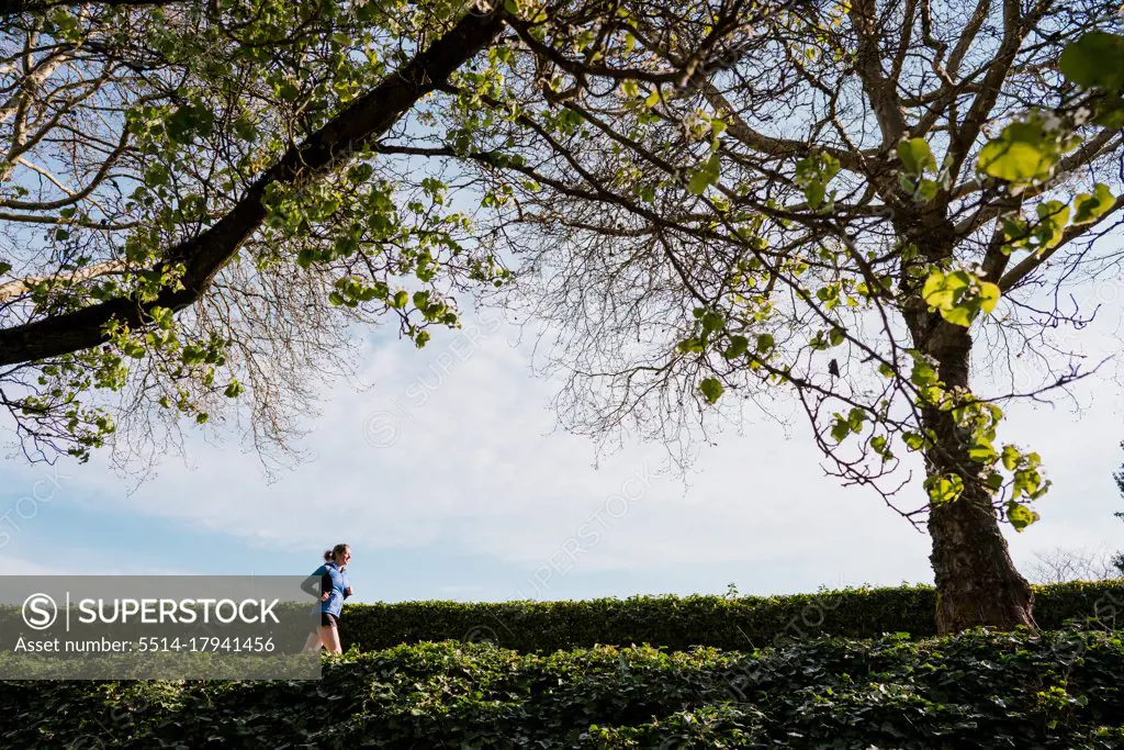 Young woman runs underneath tall green trees and blue skies