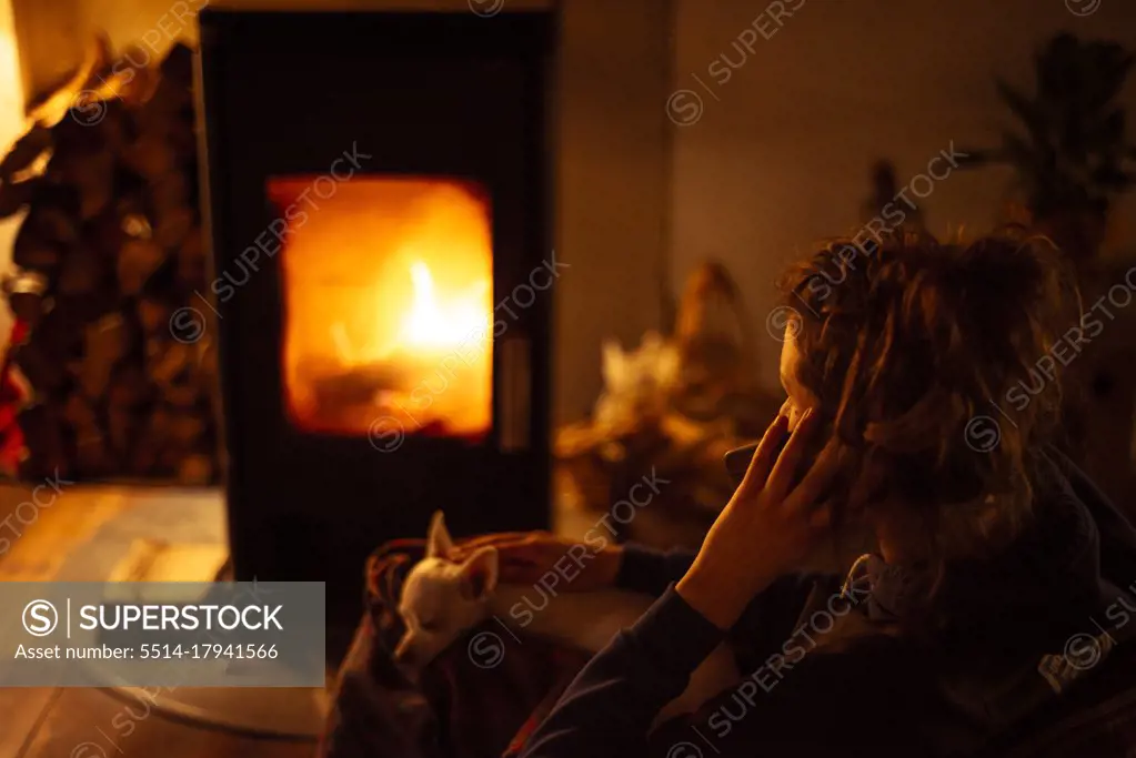 Young woman talks on phone at home by warm fire with small dog on lap