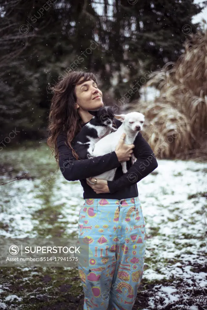 happy woman throughs head back in snow holding two small dogs in yard