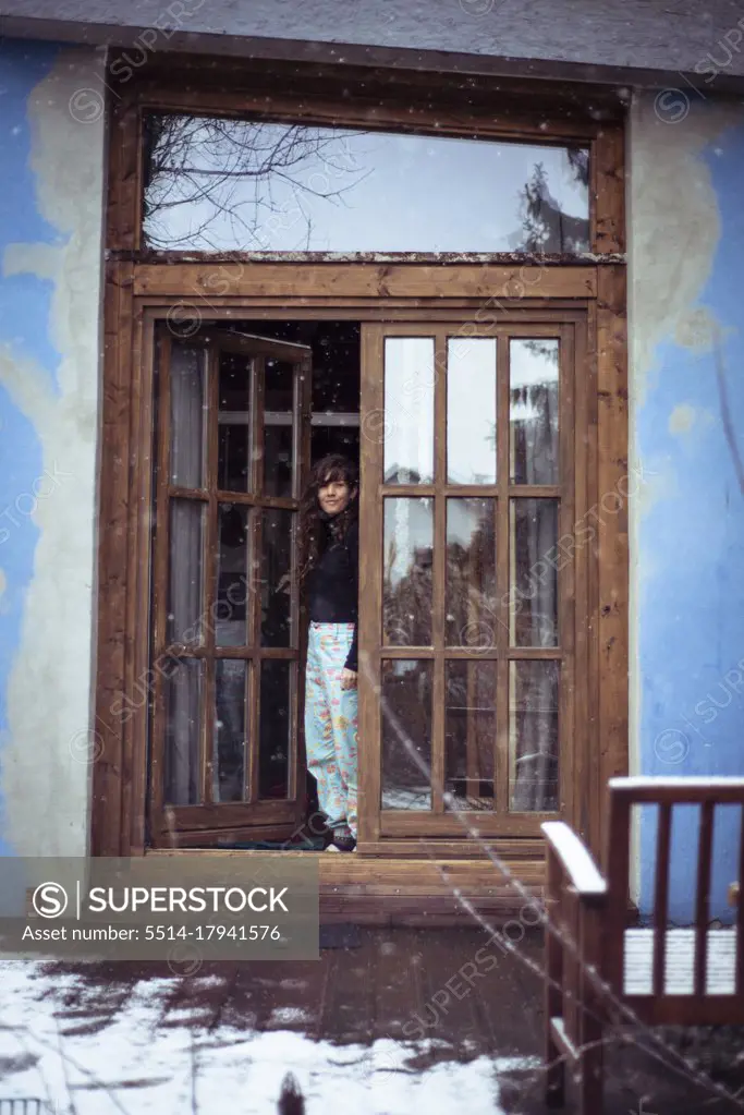 Woman with long curly hair & bright blue pants stands at wood doorway