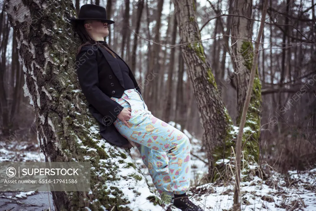 fashionable androgynous person with hat over eyes leans on snowy tree