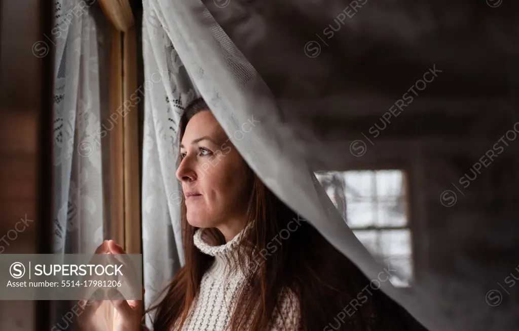 Attractive woman looking through window behind a lace curatin.