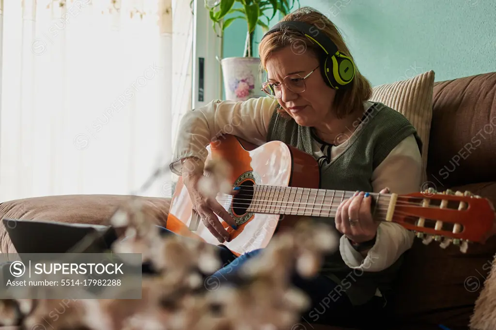Middle-aged woman with a headphones plays guitar on the sofa at home