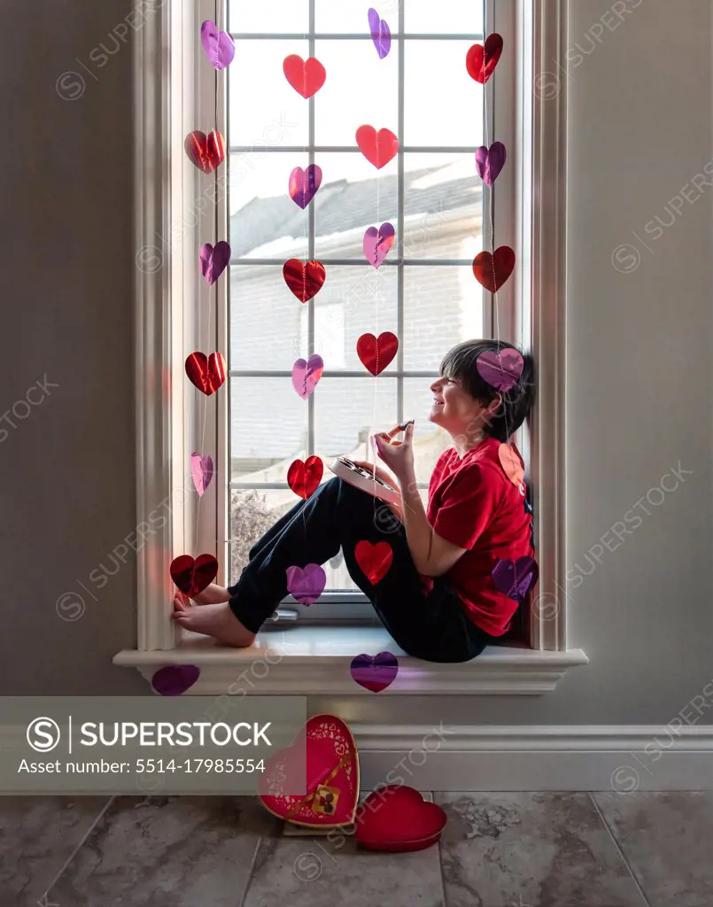Boy eating chocolates in window decorated with hearts for Valentines.