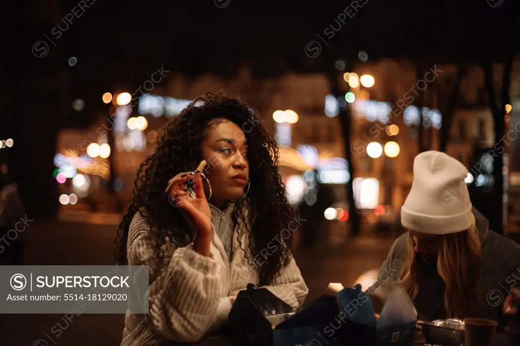 Young women eating street food standing by table in city at night